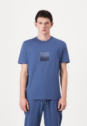 Blue cotton t-shirt featuring a square logo in darker shades with the text "HUGO" centered. Short sleeves and crew neck design.