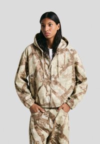 Hooded zipjacka i beige med bruna bladmönster. Har elastiska ärmslut och en avslappnad passform, tillverkad av texturerad tyg.