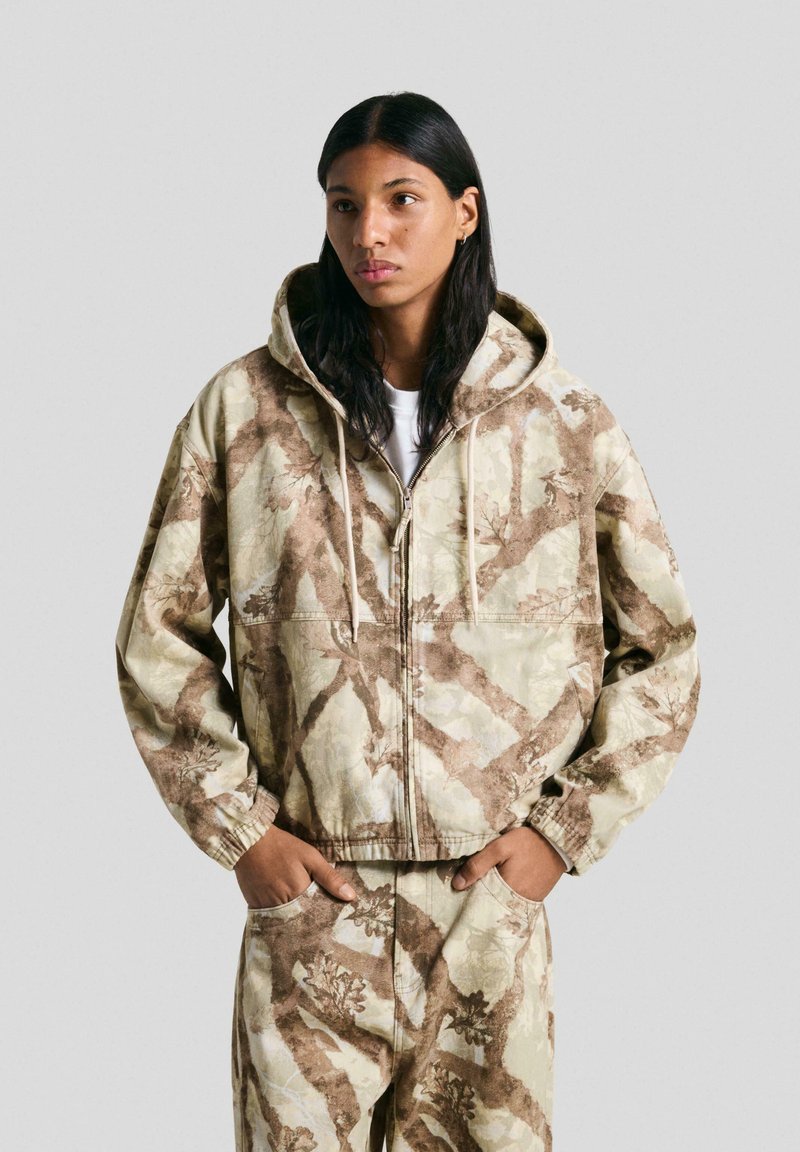 Hooded zipjacka i beige med bruna bladmönster. Har elastiska ärmslut och en avslappnad passform, tillverkad av texturerad tyg.