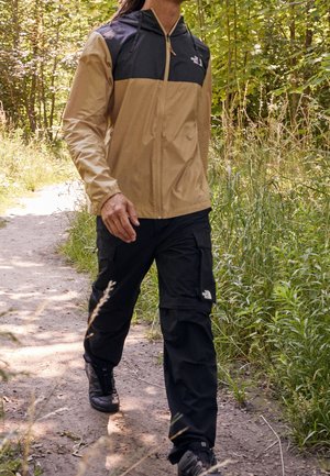 Person trägt eine beige-schwarze Jacke und schwarze Cargohosen und geht auf einem sonnenbeschienenen Erdweg umgeben von grünen Pflanzen und Bäumen.