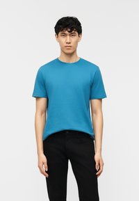 Νεαρός άνδρας που στέκεται με το πρόσωπο προς τα εμπρός, φορώντας απλό μπλε κοντομάνικο T-shirt και μαύρο παντελόνι, μπροστά σε απλό φωτεινό φόντο.