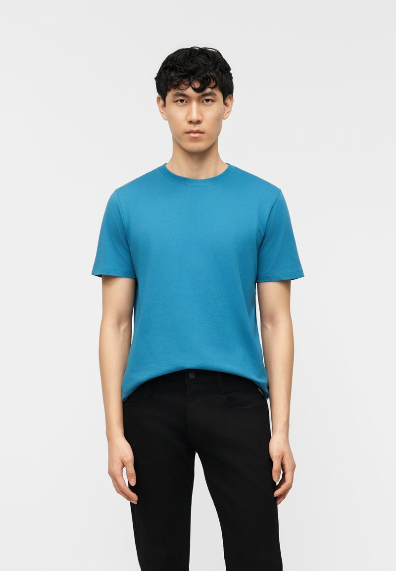 Νεαρός άνδρας που στέκεται με το πρόσωπο προς τα εμπρός, φορώντας απλό μπλε κοντομάνικο T-shirt και μαύρο παντελόνι, μπροστά σε απλό φωτεινό φόντο.