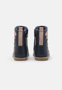 Ein Paar marineblaue Kinderstiefel mit beigen Sohlen, glitzernden pinken Rückstreifen und seitlichen Reißverschlüssen, von hinten auf einem weißen Hintergrund gezeigt.