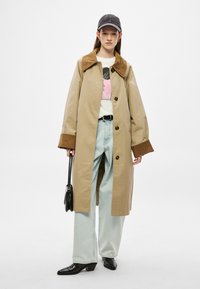 Beež trentskulptuur pruuni krae ja mansettidega, nööpidega, kantud graafilise t-särgi ja helesiniste teksade peal. Must käekott ja kontsaga saapad.