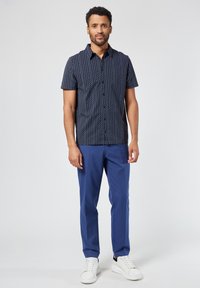 Lyhythihainen tummansininen paita, jossa on geometrinen kuvio, yhdistetty sinisiin housuihin ja valkoisiin tennareihin. Paidassa on nappikaulus.