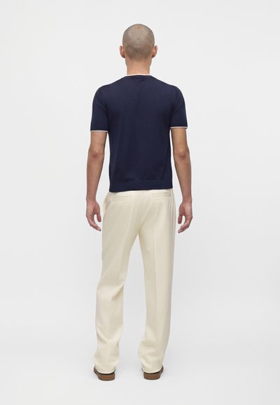 Homme à la tête rasée, debout de dos, portant une chemise bleu marine à manches courtes, un pantalon crème et des chaussures marron sur fond uni.
