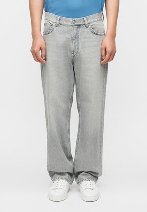 Osoba ubrana w luźne, jasnoszare dżinsy i białe tenisówki, stojąca na jednolitym tle z rękami swobodnie opuszczonymi wzdłuż ciała.