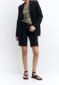 Schwarze, taillierte Blazerjacke über einem grünen gemusterten Oberteil, kombiniert mit schwarzen Jeans-Shorts. Das Model trägt schwarze flache Sandalen mit Riemen.
