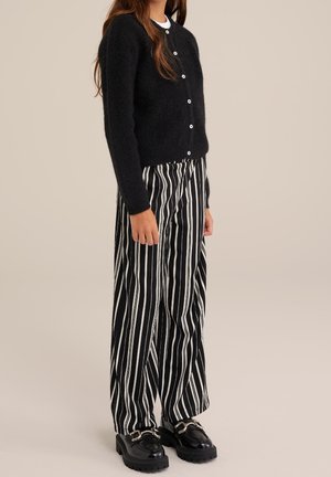 Persoon draagt een zwart gebreid vest met knopen, zwart-witte verticale gestreepte wijde broekspijpen en zwarte loafers met zilveren gespen.