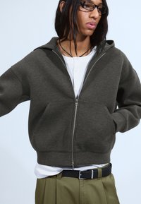 Czarny szary hoodie na zamek wykonany z miękkiego materiału, z przedni�ą kieszenią, w oversize'owym kroju i z kapturem, noszony na białej koszuli z guzikami.