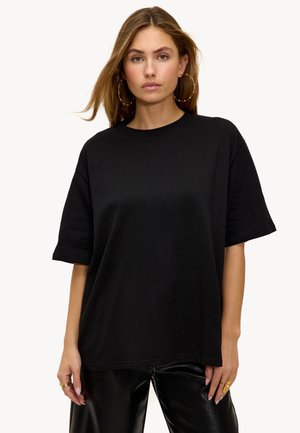 Mujer con cabello largo que lleva grandes pendientes de aro, una camiseta negra holgada y pantalones de cuero negros, de pie contra un fondo claro y sencillo.