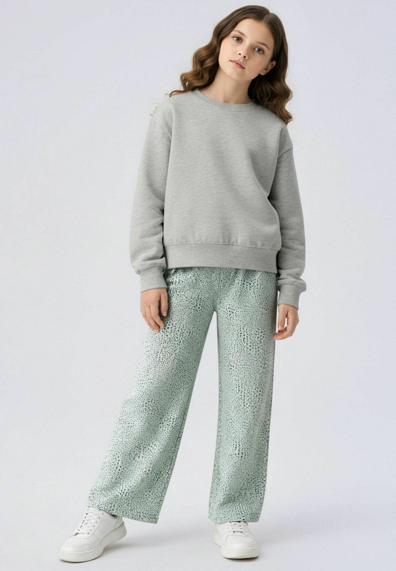 Fille aux cheveux ondulés portant un sweat-shirt gris, un pantalon ample à motifs vert clair, et des baskets blanches, debout sur un fond blanc uni.