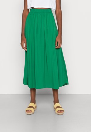 Frau trägt einen hoch taillierten, plissierten grünen Midirock mit einem weißen Cropped-Top und gelben offenen Sandalen vor einem einfarbigen Hintergrund.