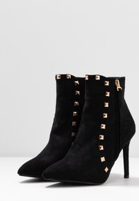 Bottes chevilles en daim noir avec bouts pointus, clous pyramides dorés sur les côtés et un talon aiguille audacieux. Fermeture éclair sur un côté.