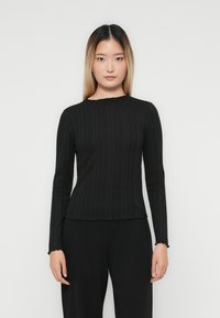Tânără cu păr drept de la negru la blond ombre, purtând un top lung cu manșon ribbed negru și pantaloni negri, pe un fundal alb simplu.