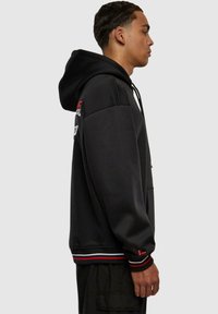 Schwarzer Hoodie mit einer Fronttasche, Kapuze mit Kordelzug und kontrastierenden gerippten Bündchen und Saum mit roten und weißen Streifen. Seitenansicht.