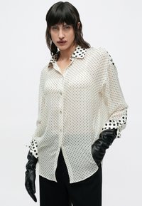 Cămașă crem transparentă cu buline negre, guler contrastant cu buline și accent negru pe mâneci lungi, asortată cu pantaloni și mănuși negre.