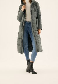 Długa, pikowana kurtka w kolorze oliwkowej zieleni z kapturem, z zamkiem błyskawicznym z przodu i kieszeniami po bokach. Zestawiona z niebieskimi obcisłymi dżinsami i czarnymi botkami na obcasie.