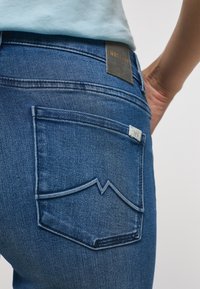 Sötét kék farmernadrág, jellegzetes ferde hátsó zsebfelülettel és "MUSTANG" felirattal ellátott bőr címkével a derékrészen.