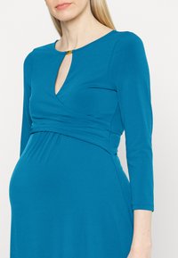 Vestido de maternidade em teal com mangas três quartos, apresentando um decote em gota e um design de corpete cruzado, feito de tecido elástico e suave.