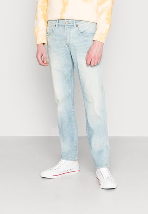 Homme portant un jean bleu clair délavé, des baskets blanches montantes avec une bande rouge, et un sweat jaune tie-dye debout sur un sol gris.