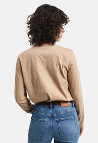Langärmeliges beiges Baumwollhemd, getragen mit blauen Jeans. Das Hemd hat ein einfaches Design, einen runden Ausschnitt und einen leicht lockeren Schnitt.