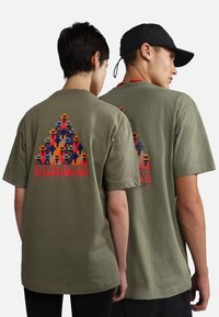 Camisetas de algodón verde oliva con un gráfico colorido de personas con sombreros en la parte posterior, con el logo "NAPAPIJRI" en rojo debajo.