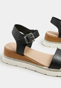 Czarne sandały na kostkę z ekoskóry na drewnianym koturnie, z przodu ozdobione cyrkoniami i wyposażone w wyściełaną wkładkę.