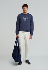 Hanorac bleumarin cu text alb, pantaloni deschiși, și geantă bleumarin. Adidași gri și croială relaxată. Design minimal, fără modele vizibile.