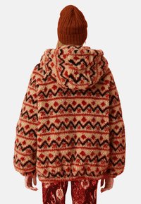 Flauschige Kapuzenjacke mit einem geometrischen Muster in Rot, Schwarz und Creme. Sie hat übergroße Ärmel und eine weiche Textur. Strickmütze in Braun.