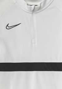 Top blanco de Nike con una franja horizontal negra, un cierre de cuarto en el cuello y un pequeño logo negro en el lado izquierdo del pecho. Textura suave.