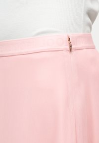 Κοντινό πλάνο της μέσης μιας ανοιχτό ροζ φούστας με διακριτικό υφασμένο λογότυπο και μικρό χρυσό φερμουάρ, επάνω σε λευκό φόντο κορυφής.