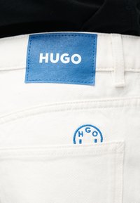 Λευκά τζιν από τζιν ύφασμα με μπλε ορθογώνια ετικέτα μάρκας "HUGO" στη μέση και κυκλικό ραμμένο λογότυπο πάνω από την πίσω τσέπη.