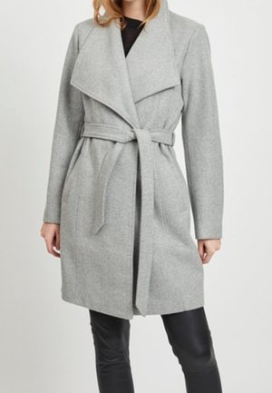 Femme portant un manteau ceinturé gris clair longueur genoux avec de larges revers, sur un pantalon noir et un haut noir, sur fond blanc uni.
