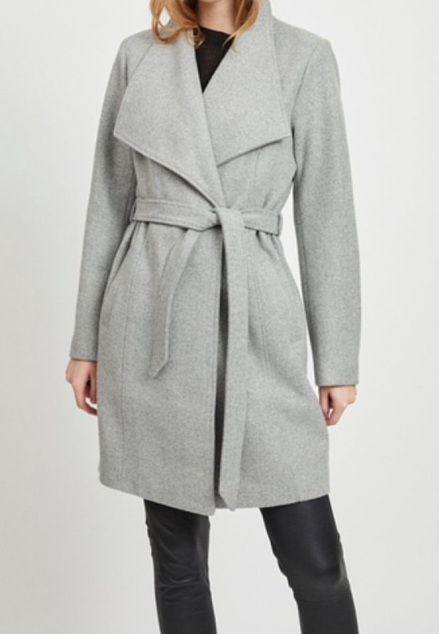 Femme portant un manteau ceinturé gris clair longueur genoux avec de larges revers, sur un pantalon noir et un haut noir, sur fond blanc uni.
