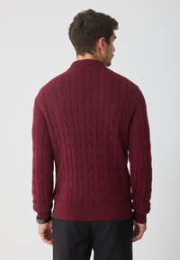 Bordowy sweter z splotem w warkocze, z wysokim kołnierzem, ściągaczami przy rękawach i u dołu; z teksturowanym, tkanym wzorem, który dodaje głębi.