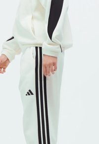 Bijela trenirka s crnim detaljima i tri pruge duž hlača; ima ležeran kroj i rebraste manšete. Adidas logo na nozi.