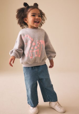 Lachende peuter met krullend haar, die een grijze trui met een roze paardenontwerp, losse blauwe jeans en witte sneakers draagt.