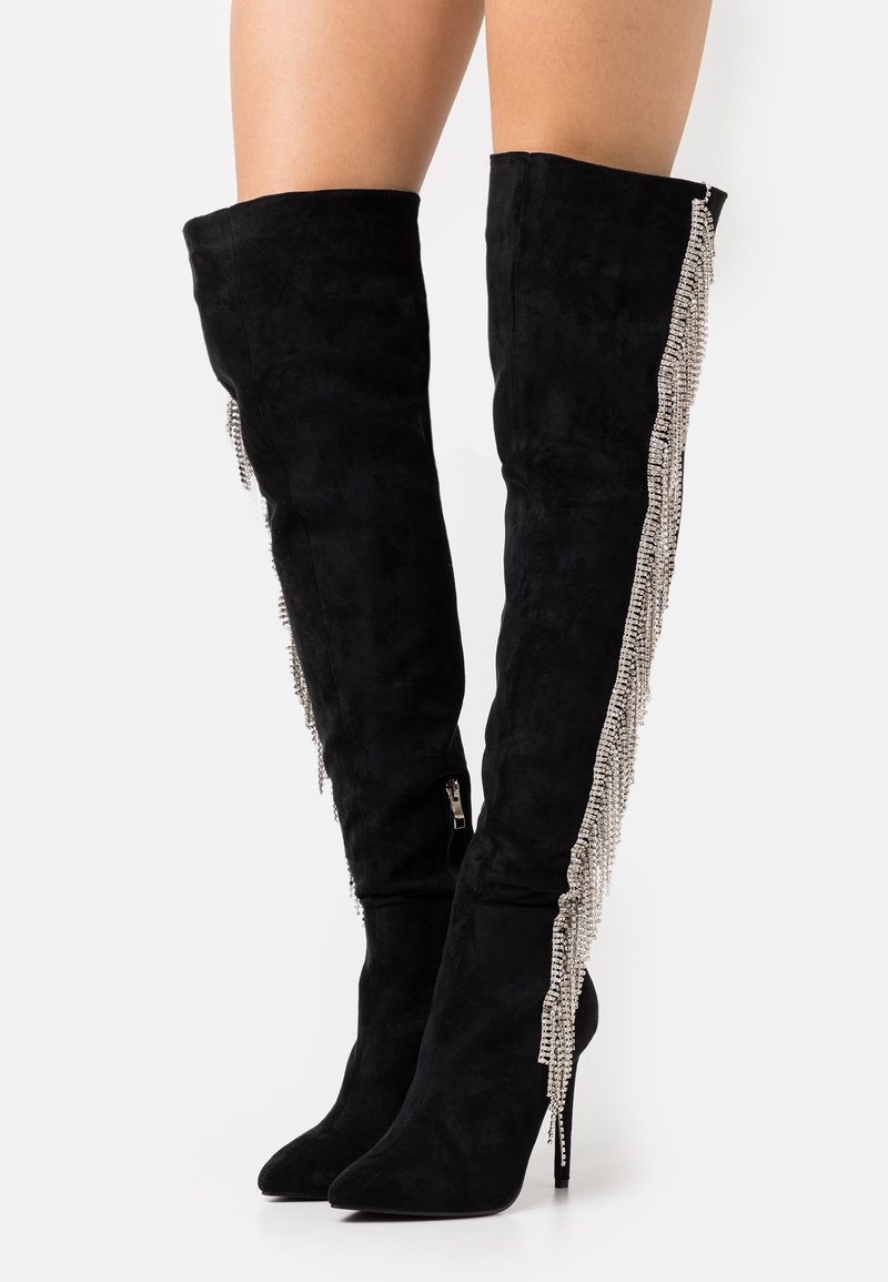 Botas de ante negro hasta el muslo con puntera afilada y tacones de aguja, con un detalle de cadena plateada a lo largo de los lados exteriores.