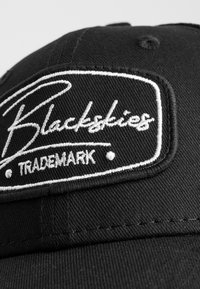 Schwarze Baseballmütze aus Baumwolltwill mit einem weißen gestickten Logo, das "Blackskies" und "TRADEMARK" auf einem rechteckigen Patch zeigt.