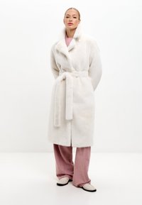 Vit faux fur-kappa med sjalkrage, knytbart midjeband och en enkel knappdetalj, bärs över ljusrosa byxor och skor. Slät textur.