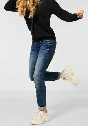 Person trägt dunkelblaue Jeans, schwarzen, geknöpften Pullover und weiße Turnschuhe, posiert mit einem angehobenen und angewinkelten Bein.