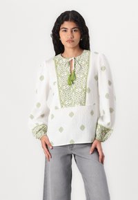 Hvit langermet bluse med grønn blomsterbroderi, nøkkelhulls-utskjæring med dusk og mønstrede mansjetter; lett og teksturert stoff.