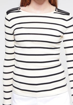 Femme portant une chemise blanche à manches longues avec des rayures horizontales noires et des détails à fermeture éclair sur les deux épaules, associée à un pantalon blanc.