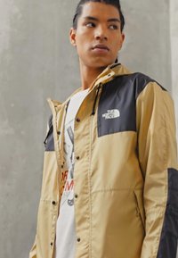 Lagana jakna s blokovima boja, smeđe boje tela i crnim rukavima. Ima kapuljaču, dugmad za snap i logo The North Face na prsima.