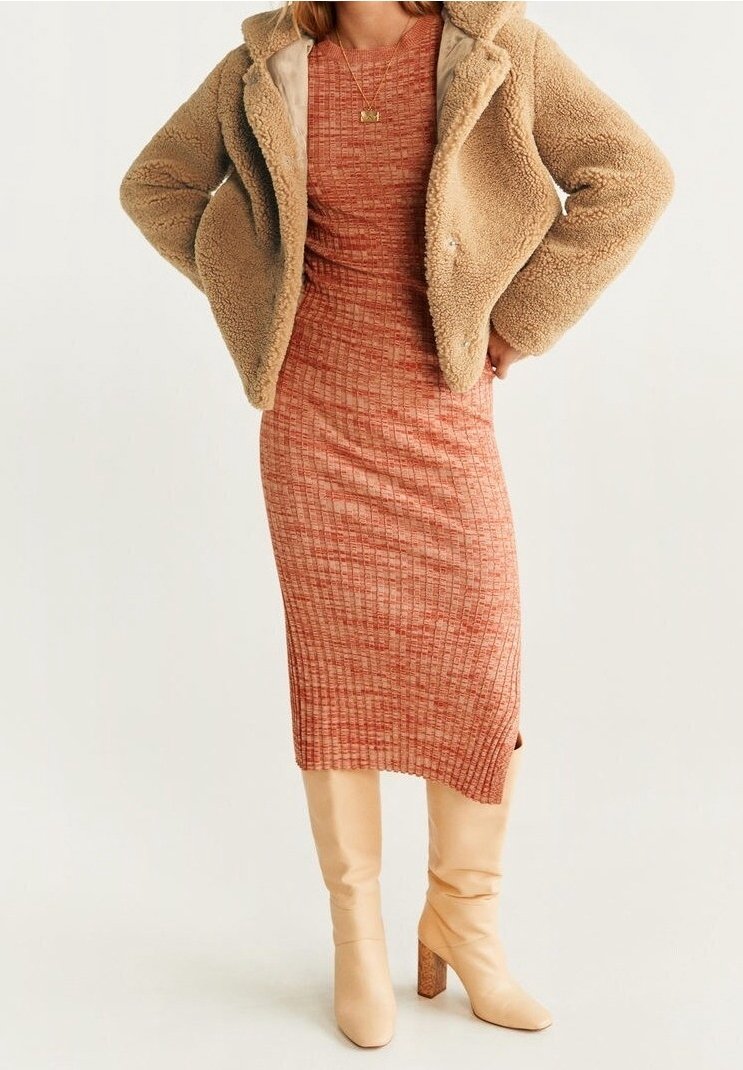 Femme portant une robe midi ajustée couleur rouille, une veste teddy beige, des bottes beiges jusqu'aux genoux et un collier en or, se tenant les mains sur les hanches.