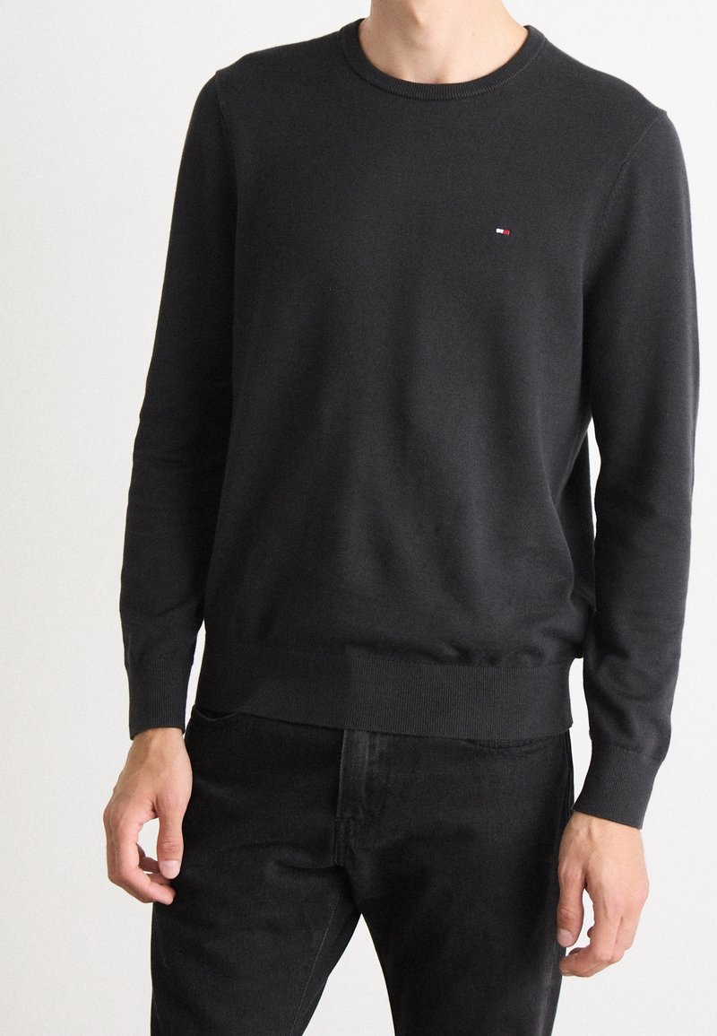 Homme portant un pull noir ajusté à col rond avec des manches longues et un petit logo rouge, blanc et bleu sur le côté gauche de la poitrine.