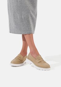 Mocassini in pelle beige con due strap decorative, suola piatta in gomma bianca, punta rotonda e superficie texturizzata. Indossati con una gonna grigia in maglia.