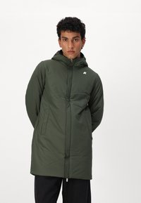 Veste verte isolée avec capuche, fermeture éclair à l'avant et deux poches latérales. Présente un logo sur la poitrine gauche et une texture lisse.