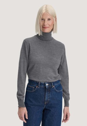 Mature woman with straight white hair wearing a gray turtleneck sweater and blue high-waisted jeans against a neutral background.